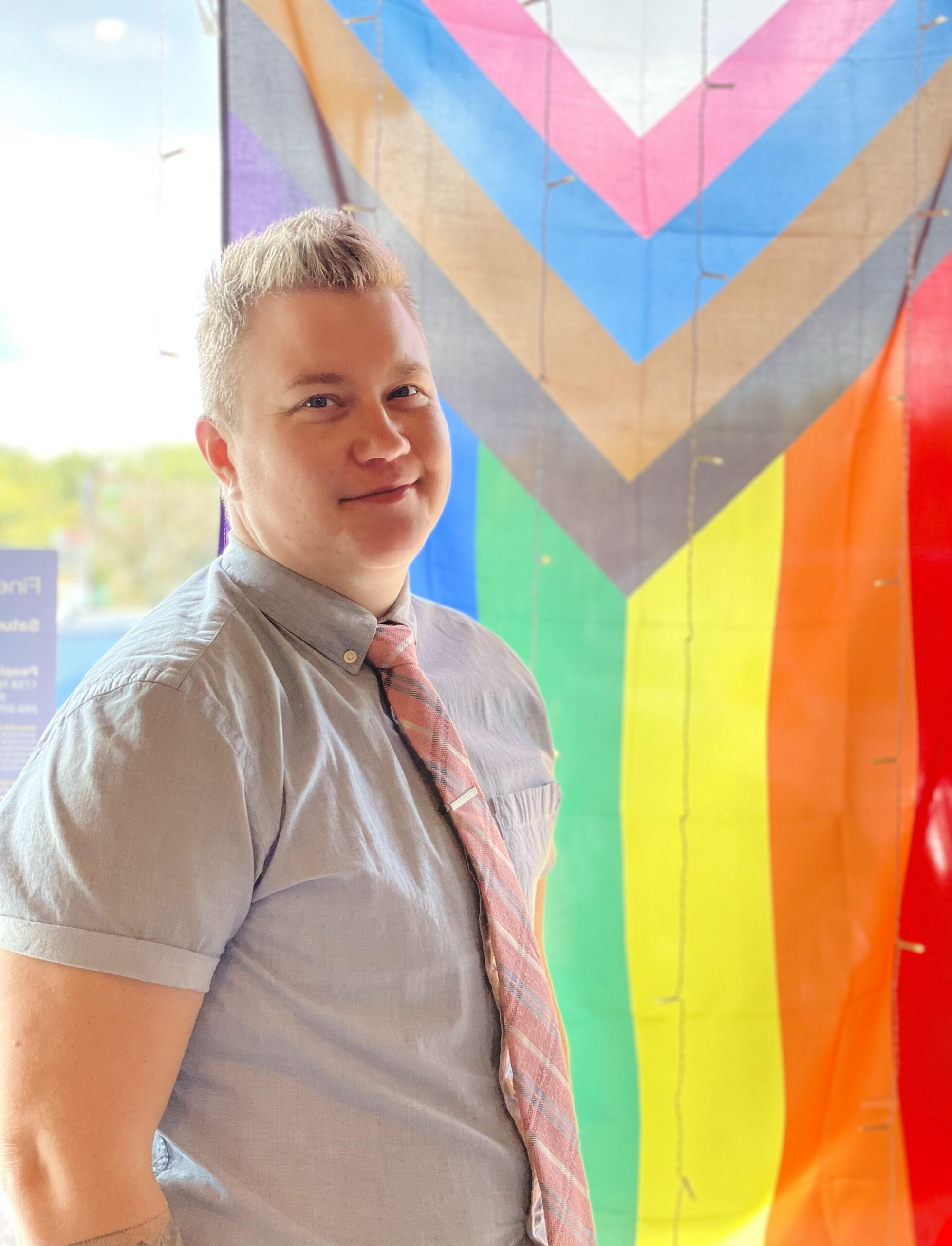 A person wearing a short sleeve shirt and pink necktie stands in front of a window with a Progress Pride Flag hanging in it.